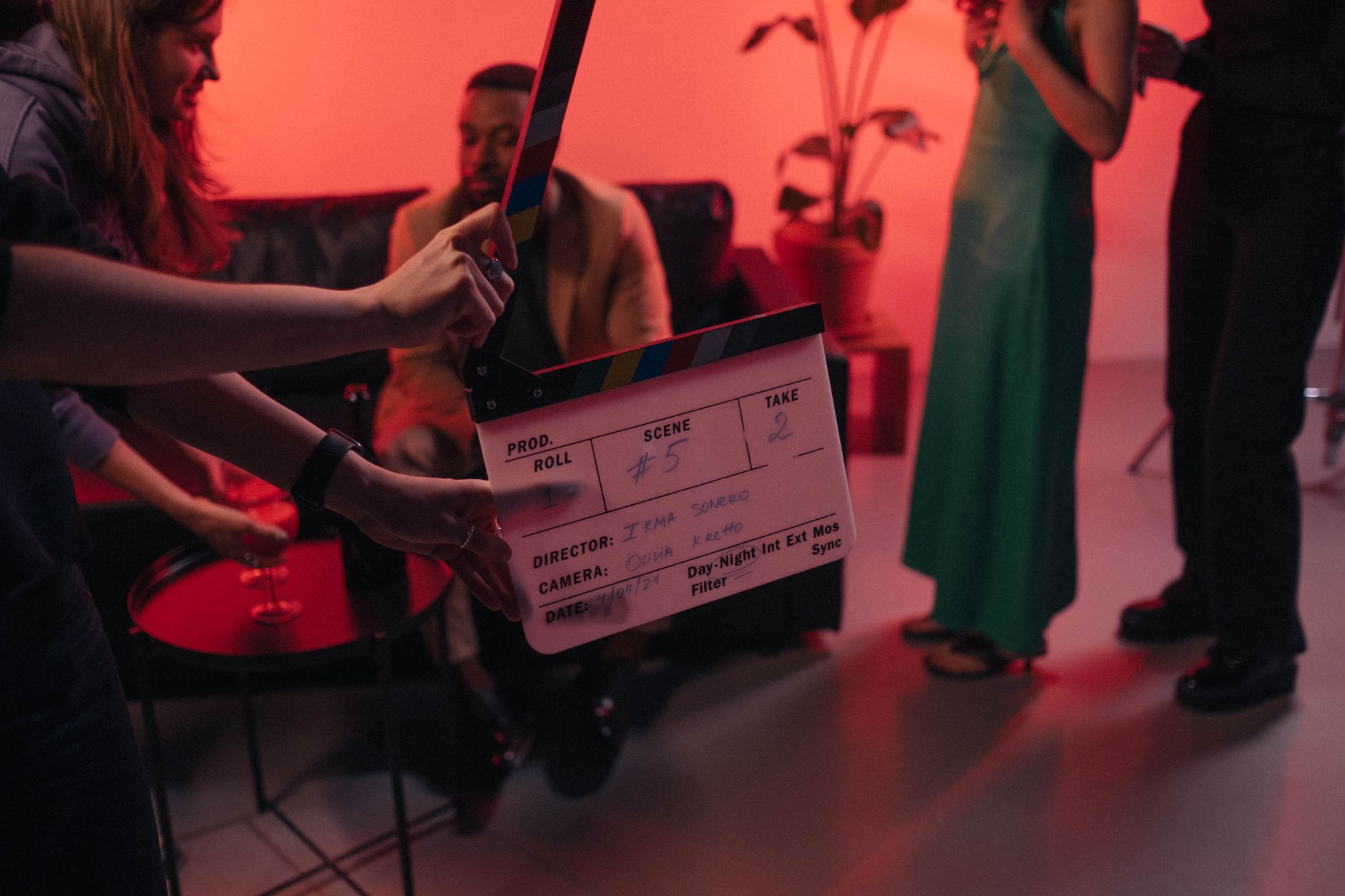 Des mains tiennent une claquette de cinéma sur un plateau éclairé de rouge, avec des personnes à l’arrière-plan.