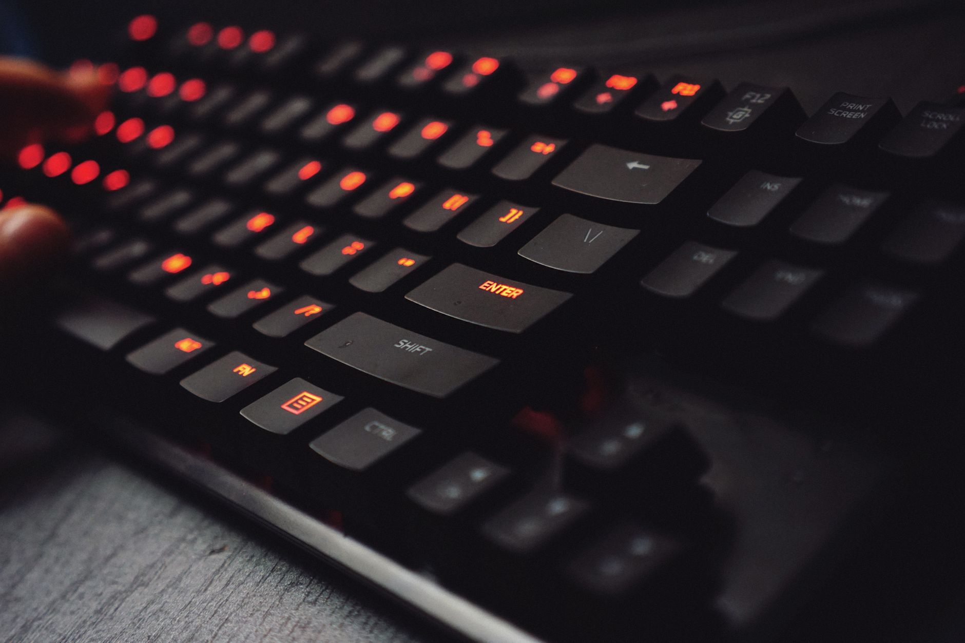Gros plan sur un clavier mécanique noir compact aux touches rétroéclairées en rouge, avec des doigts flous à gauche, posé sur un bureau gris.