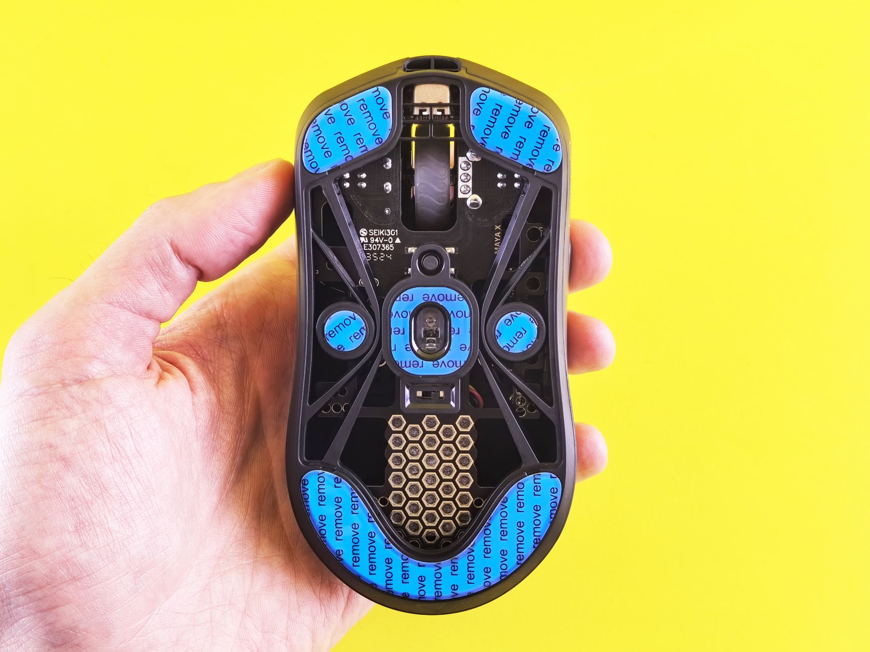 Main tenant une souris de jeu noire retournée, dessous ajouré avec patins bleus et grille hexagonale, sur fond jaune.