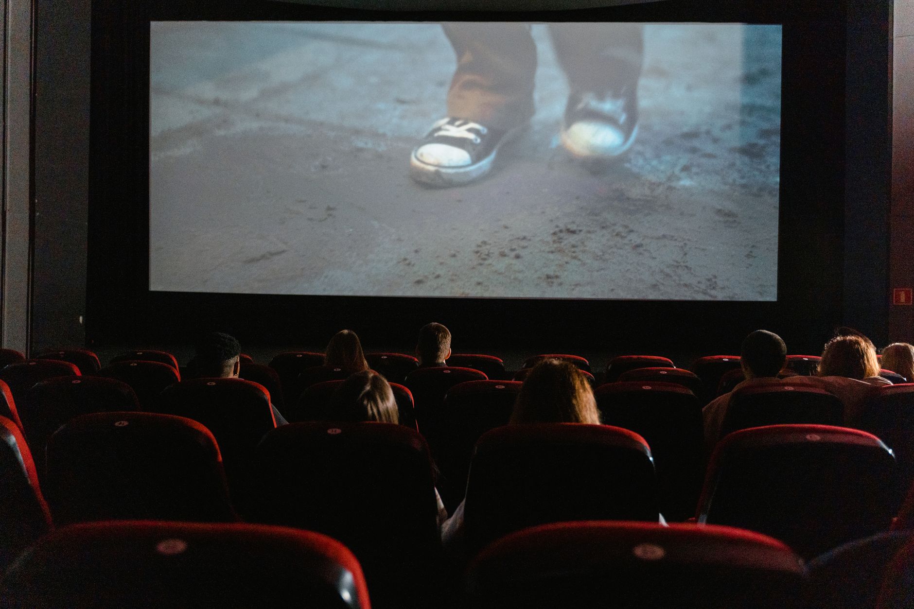 Spectateurs de dos dans une salle de cinéma, assis sur des fauteuils rouges face à un grand écran montrant des pieds en baskets.