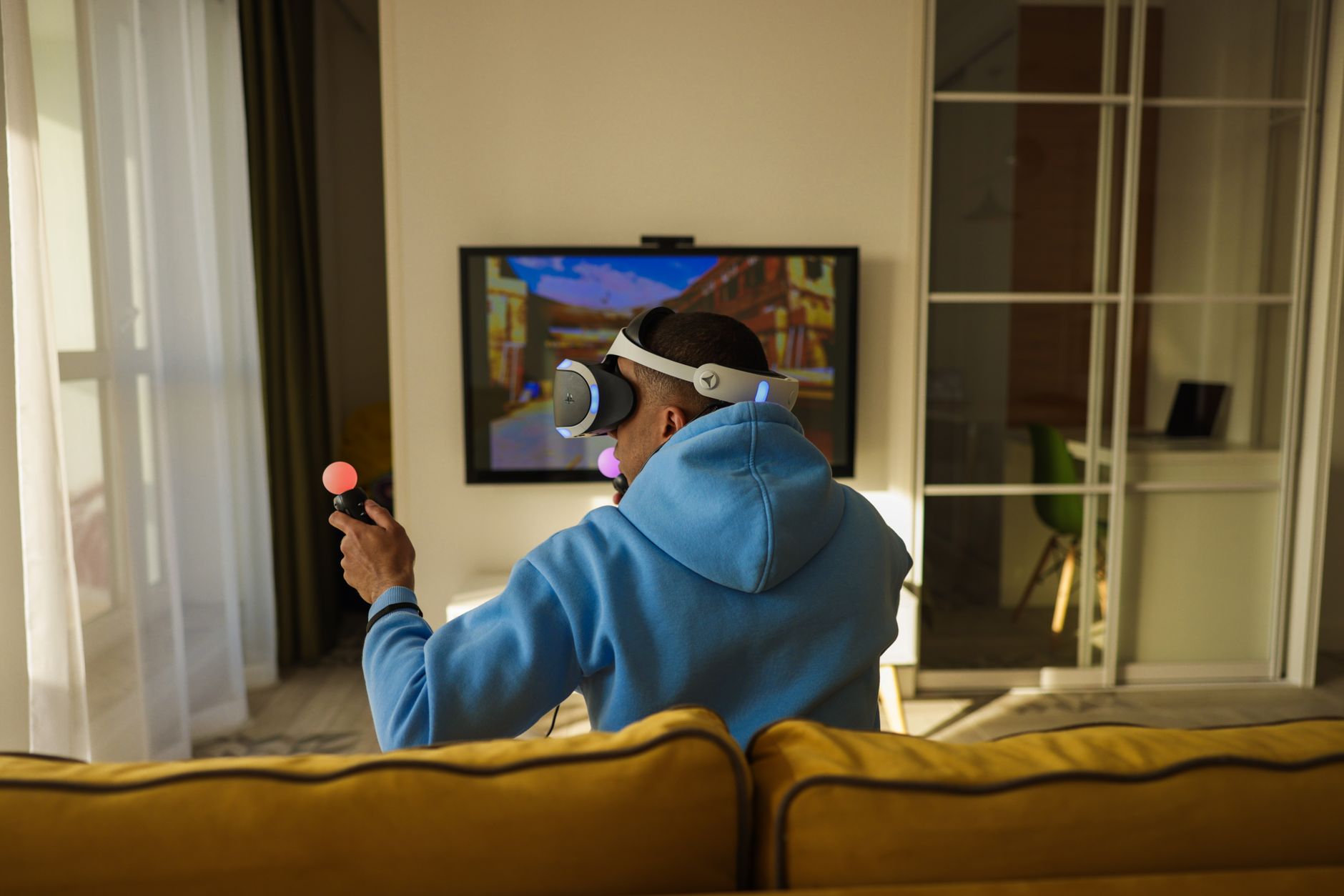 Un homme portant un casque de réalité virtuelle et des manettes lumineuses face à un écran de télévision dans un salon.