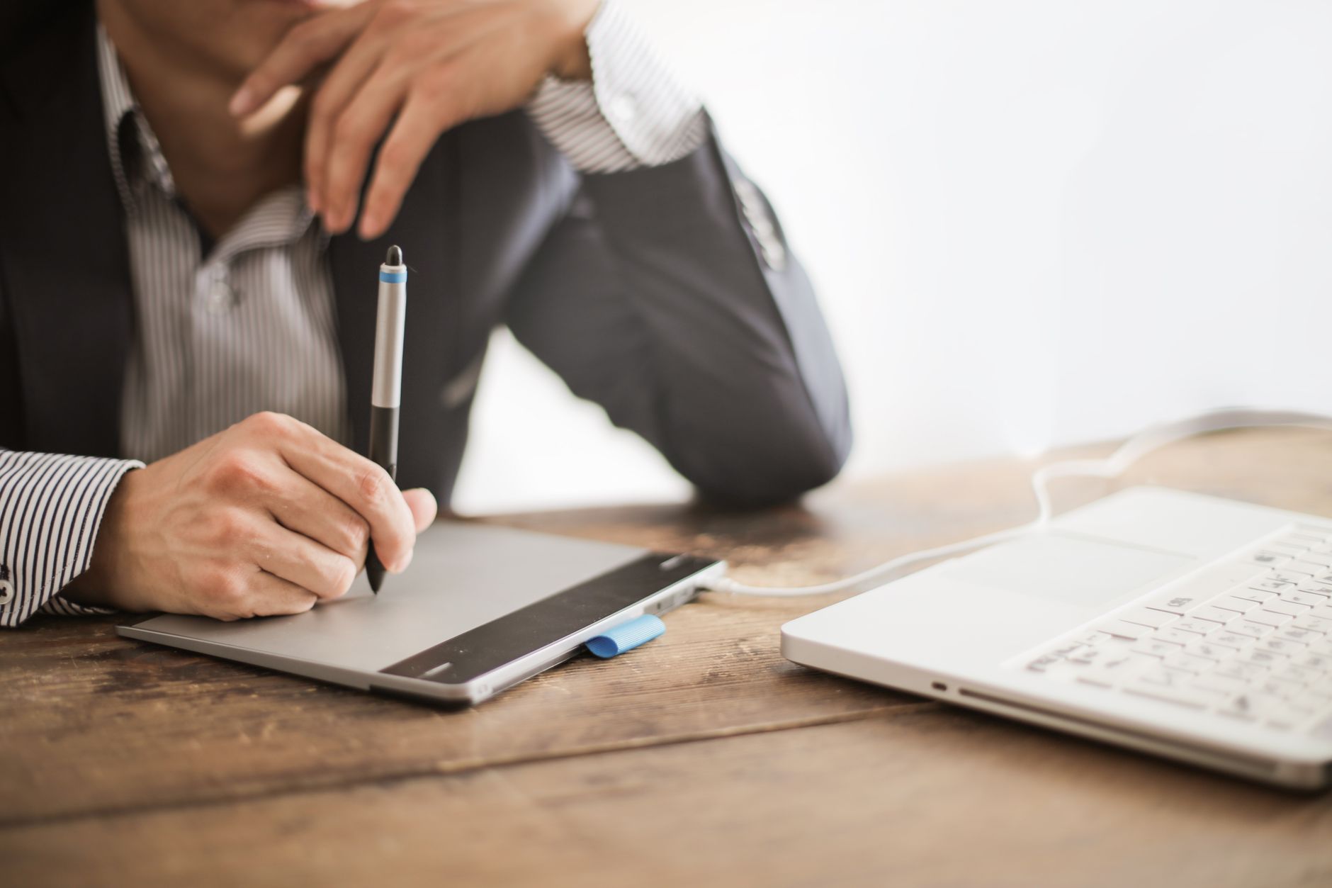 Une personne en veste dessine au stylet sur une tablette graphique posée sur un bureau en bois, à côté d’un ordinateur portable blanc.